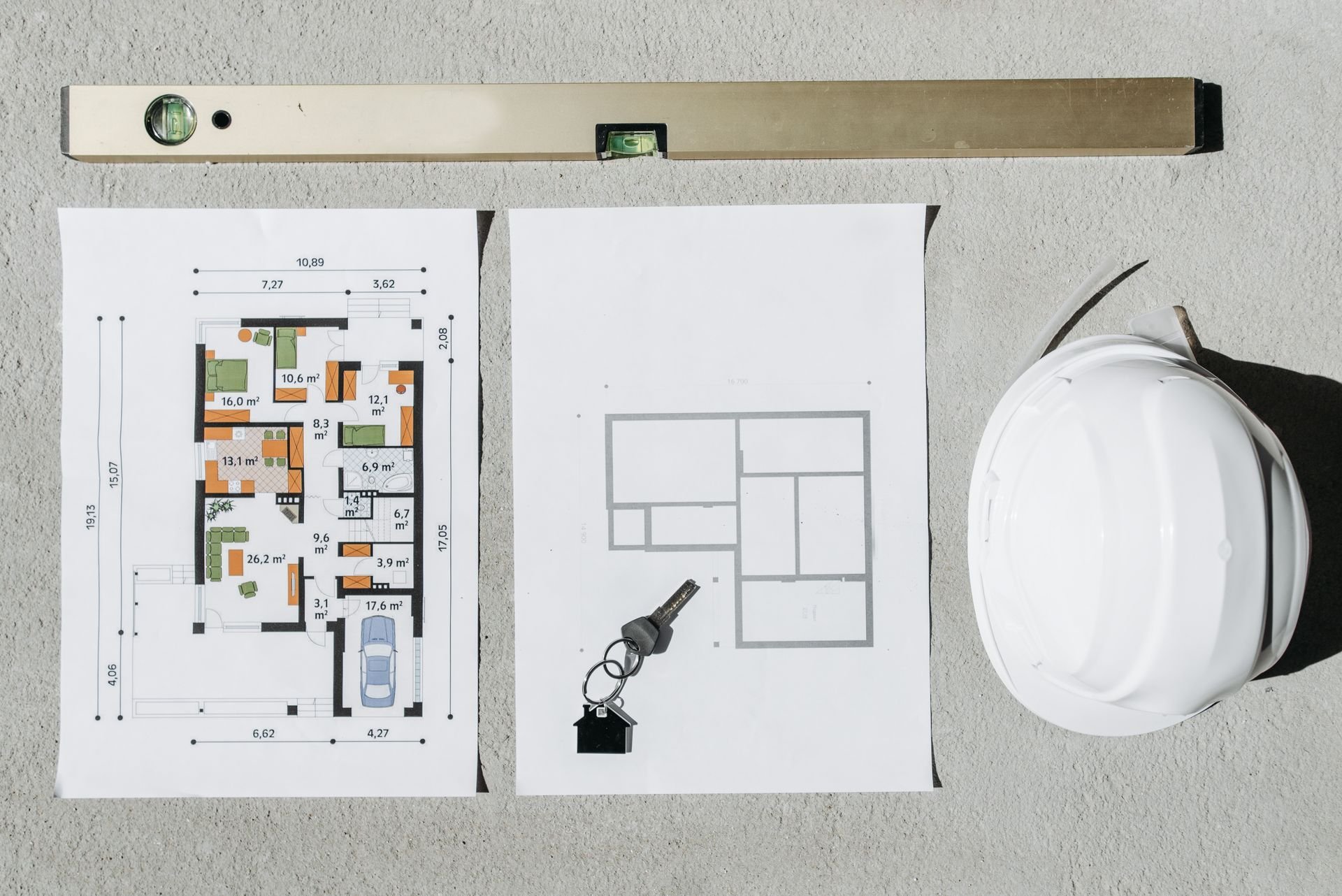 Construction plans, a spirit level, keys and a white hard hat laid out on a concrete floor at a building site