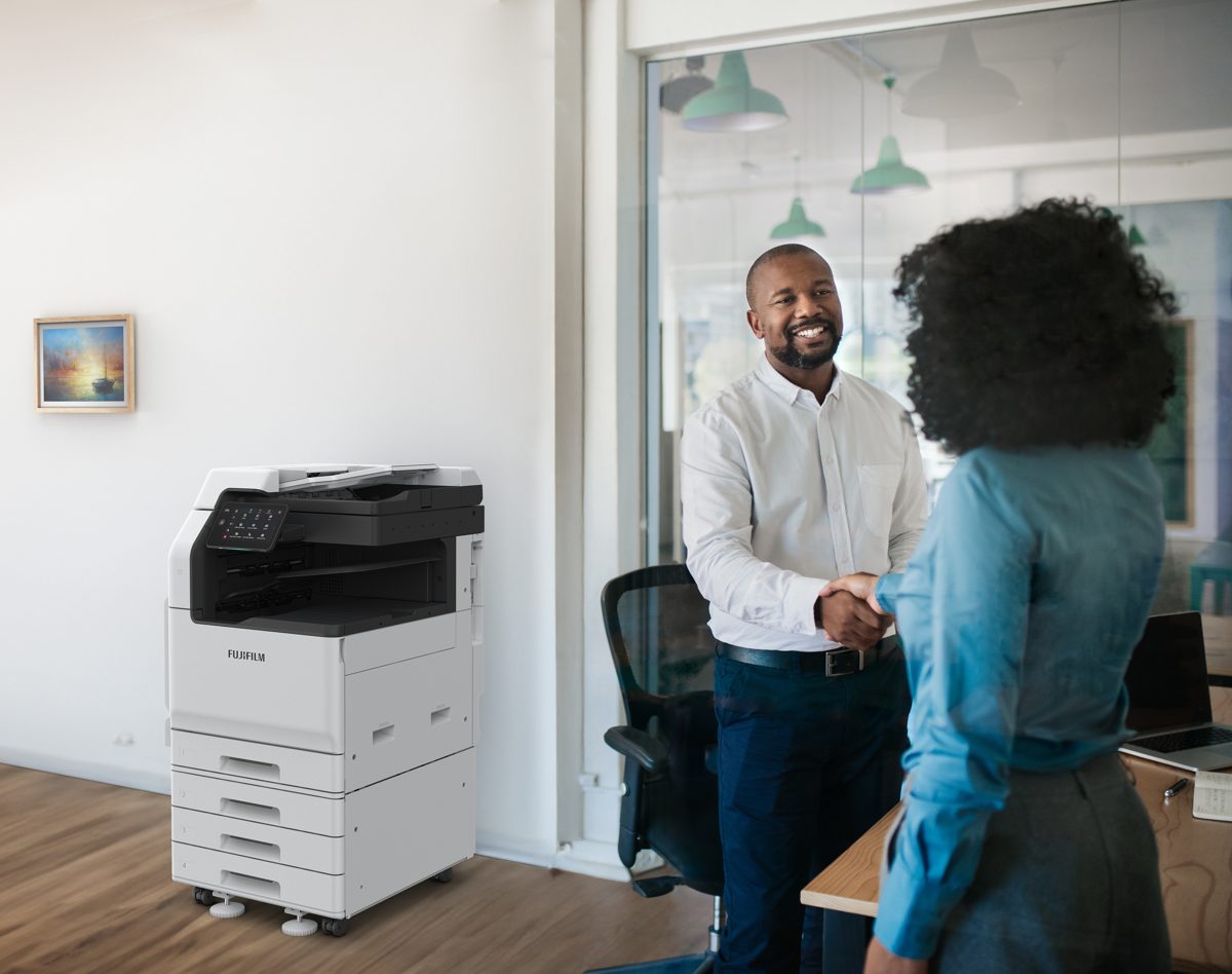 Fujifilm Apeos C3060 color multifunction printer in an office, with two colleagues shaking hands nearby